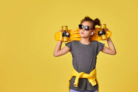 cool little child girl in sunglasses posing with yellow skateboard on yellow background. copy space