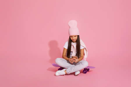 little child girl sitting on skateboard and using smartphone over pink background