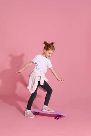 cute little child girl in casual clothes riding skateboard against pink background.の写真素材