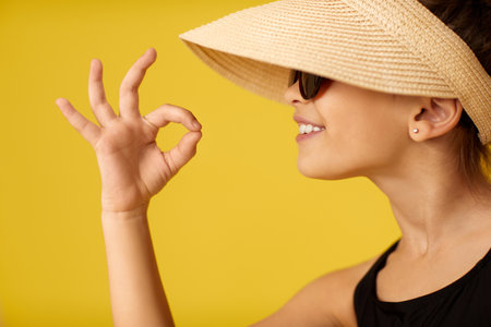 beautiful smiling little child girl in cap showing Ok sign on yellow background.の写真素材