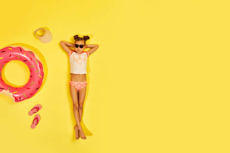 little child girl in swimwear and sunglasses lying on yellow background with inflatable rubber ring. Top view. copy spaceの写真素材
