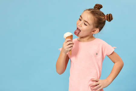 happy little girl eating vanilla ice cream in a waffle cone on blue background. copy space for textの写真素材