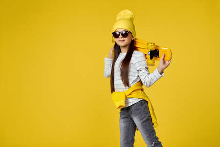 cool hipster little child girl in sunglasses posing with yellow skateboard on studio background....
