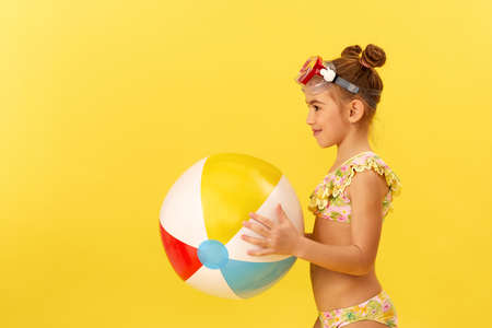 adorable little child girl in swimwear and swimming glasses with beach ball on yellow background. side viewの写真素材
