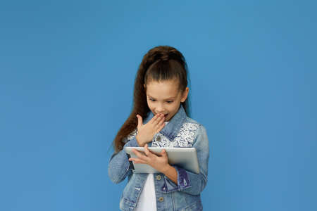 Surprised little child girl in denim using digital tablet on blue background