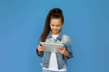pretty smiling little child girl in denim using digital tablet on blue backgroundの写真素材