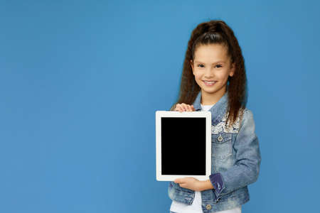 adorable smiling little child girl shows empty tablet on blue background. copy spaceの写真素材