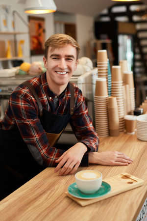 happy male waiter in apron holding cup of hot drinkの写真素材