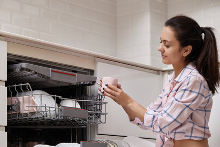 attractive woman unloading cup from open automatic built-in dishwasher machine with clean utensils inside in modern white kitchen.の写真素材