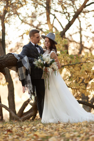 beautiful happy stylish bride in hat and groom standing outdoor in autumnの写真素材
