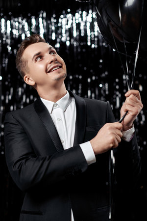 Happy handsome man in black jacket holding black air balloons on glitter background.の写真素材