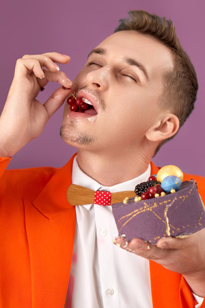 handsome birthday man in orange jacket eating cake on purple backgroundの写真素材