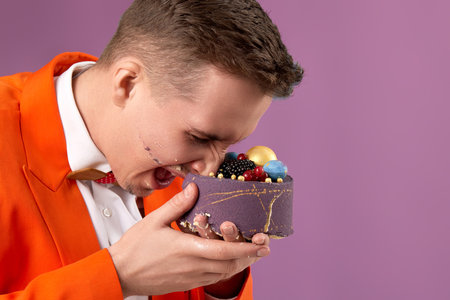 birthday man in orange jacket bites cake on purple backgroundの写真素材