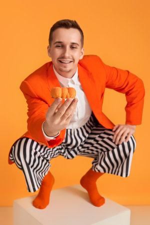 extravagant funny young man in orange jacket and striped pants holding cupcake on orange background.の写真素材