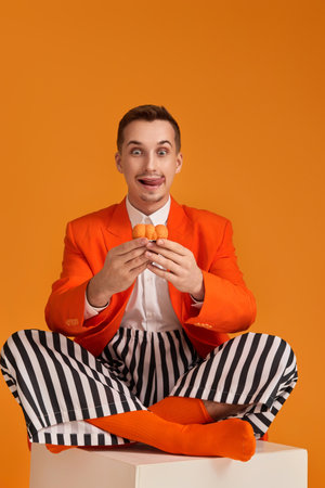extravagant funny young man in orange jacket and striped pants holding cupcake on orange background.の写真素材