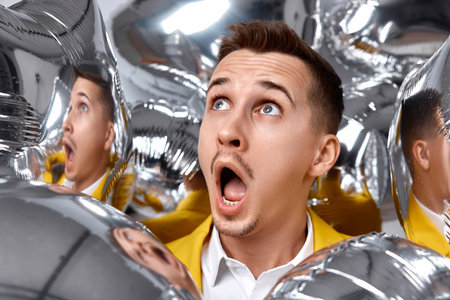 portrait of amazed man in yellow jacket with a lot of silver air balloons at birthday partyの写真素材
