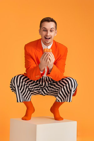extravagant funny young man in orange jacket and striped pants holding cupcake on orange background.の写真素材