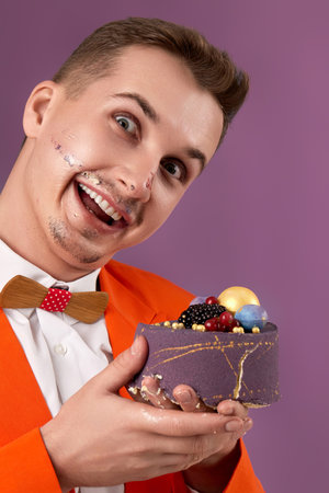 funny birthday man in orange jacket eating cake on purple backgroundの写真素材
