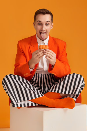 extravagant funny young man in orange jacket and striped pants holding cupcake on orange background.の写真素材
