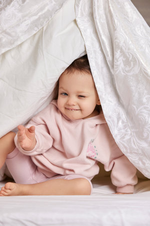 cute funny little child girl looks out from under the blanket on the bedの写真素材