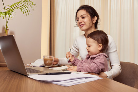 Cheerful pretty businesswoman working on laptop at home with her little child girl. mom spending time with her cute babyの写真素材