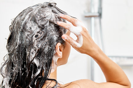 Young woman taking refreshing shower in white modern bathroom. Girl washing her hair, back viewの写真素材