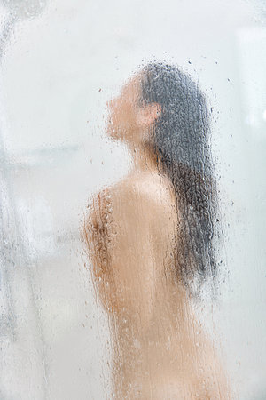 Unfocused woman washing long hair in shower behind glass with drops. Focus on dropsの写真素材