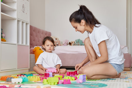 cute baby girl and her loving mom playing with toy bricks in the children room, having fun togetherの写真素材