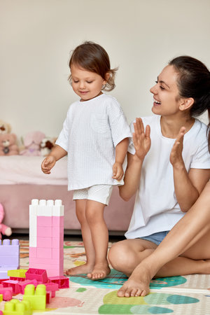 cute baby girl and her loving mother playing with toy bricks in the children room, mother applauds her child and praises herの写真素材