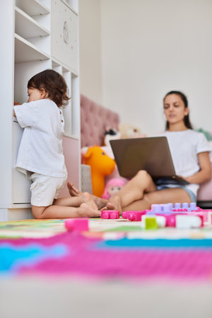 cute little baby girl playing with toys blocks and working mother sitting on floor with laptop in roomの写真素材