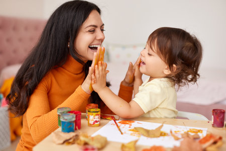 happy little child girl and mom spending good time painting hands and autumn leaves together. smiling mom and baby showing hands in yellow paintの写真素材