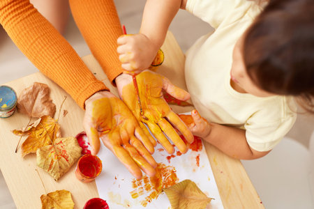 little baby girl and mother painting hands and autumn leaves together. mom and daughter showing hands in yellow paint. top viewの写真素材