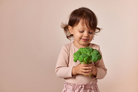 smiling child girl looking at fresh green raw broccoli on beige background. healthy baby food. kid loves vegetablesの写真素材