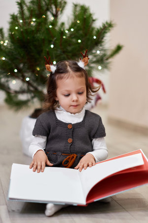 Cute little child girl reads Christmas stories and fairy tales near the Christmas tree at homeの写真素材