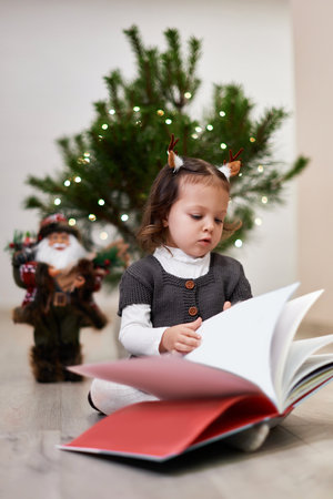 Cute little child girl reads a book near the Christmas tree at homeの写真素材