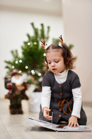 Cute little child girl reads a book near the Christmas tree at homeの写真素材
