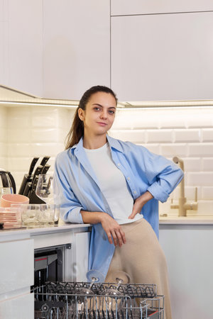 Happy woman in blue shirt standing near open dishwasher in a white modern kitchenの写真素材