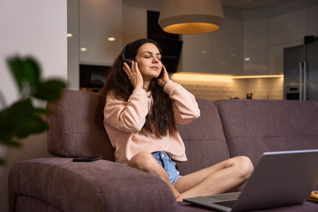 happy Caucasian woman listening to music with headphones and using laptop on couch at homeの写真素材