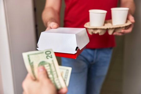 delivery man in red uniform giving food box and paper coffee cups to customer and taking payment at homeの写真素材