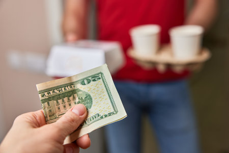 delivery man in red uniform giving food box and paper coffee cups to customer and taking payment at homeの写真素材