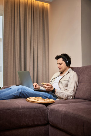 Young man sitting on sofa and eating pizza while watching movie on laptopの写真素材