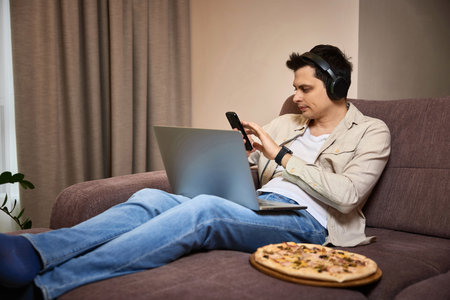 Young man sitting on sofa and eating pizza while working on laptopの写真素材