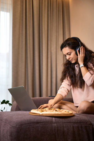 Caucasian woman working on laptop computer and eating pizza on sofa in living roomの写真素材