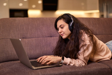 Caucasian woman in headphones using laptop and lying on sofa at homeの写真素材