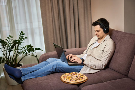 Young man sitting on sofa and eating pizza while working on laptop at homeの写真素材