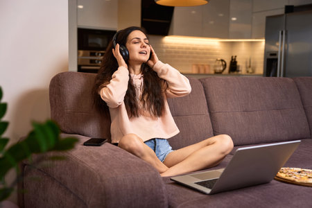 happy Caucasian woman listening to music with headphones and using laptop on couch at homeの写真素材