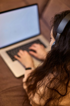 Caucasian woman in headphones typing on laptop and lying on sofa at homeの写真素材