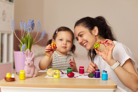 Happy mother and little daughter showing painted Easter egg at home. Mom and daughter painting eggs before Easter together.の写真素材