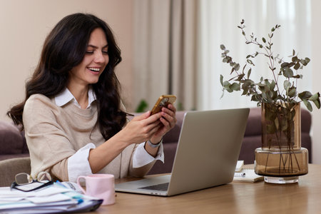 portrait of happy woman holds smart phone uses laptop in home officeの写真素材