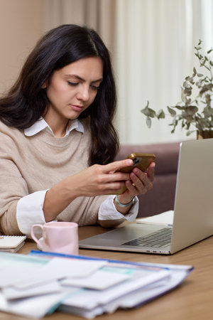 portrait of woman holds smart phone uses laptop in home officeの写真素材